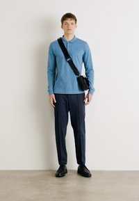 Helles blaues Langarm-Polohemd, marinefarbene Hose, schwarze Schuhe und eine kleine schwarze Tasche mit Schulterriemen, stehend vor einer weißen Wand.
