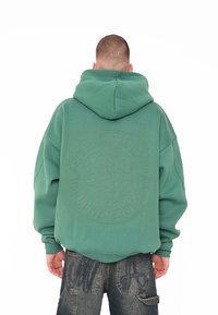 Grüne, übergroße Kapuzenjacke mit einem strukturierten, runden Logo auf dem Rücken, ausgestattet mit einer Kordelzugkapuze und gerippten Bündchen. Kombiniert mit dunklen Jeansshorts.