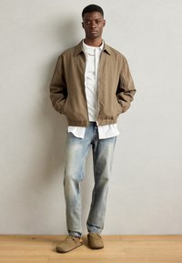Light brown bomber jacket with a collared neckline, worn over a white shirt and light blue jeans. Soft suede loafers complete the outfit.