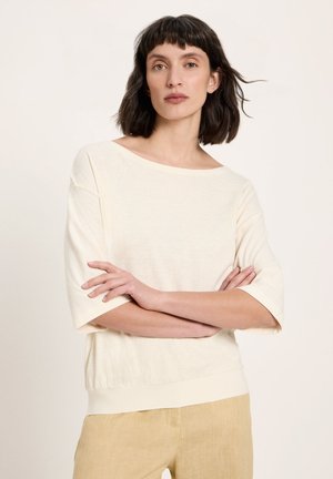 Frau mit kurzen dunklen Haaren, die ein lockeres, elfenbeinfarbenes Oberteil und beigefarbene Hosen trägt, mit verschränkten Armen vor einem einfarbigen Hintergrund stehend.