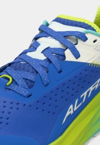 Bottine de sport bleue avec tige en mesh, accents jaunes et surface texturée. Dotée de lacets plats, de perforations pour la respirabilité et d'un logo de marque visible.