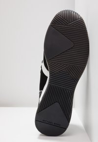 Semelle de basket noir et blanc avec motifs de grip texturés et marque "MICHAEL KORS" au talon, photographiée sur fond neutre.