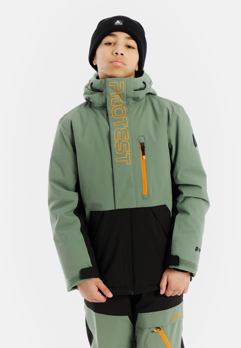 Tiener in een groen en zwarte winterjas en broek met oranje ritsen en "PROTEST" branding, en een zwarte beanie.