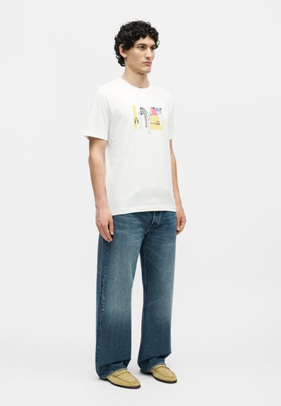 Άνδρας με κατσαρά μαλλιά, που φοράει λευκό μπλουζάκι με σχέδιο, μπλε τζιν με φαρδιά πόδια και μπεζ μοκασίνια, στέκεται μπροστά σε απλό λευκό φόντο.