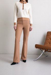 Kobieta ubrana w kremowy i brązowy sweter z guzikami, w beżowe spodnie dzwony i czarne czółenka, stoi obok brązowego skórzanego fotela.