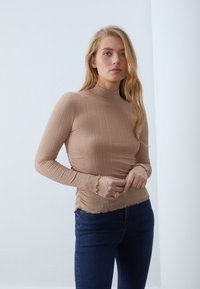 Langärmliges, beiges Rippoberteil mit hohem Kragen, gewellten Bündchen und Saum. Getragen mit dunkelblauer Jeans. Glatte Textur, figurbetontes Design.