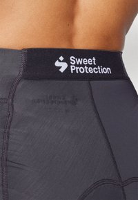 Črne atletske kratke hlače z širokim elastičnim pasom, na katerem je v belem napisan logo "Sweet Protection", gladka tekstura in brezšivne oblikovalske podrobnosti.
