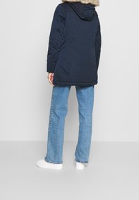 Tmavě modrý vatovaný kabát s kapucí lemovanou umělým kožíškem, s hladkým povrchem a délkou sahající po kolena, kombinovaný s světle modrými džínami širokých nohavic a bílými teniskami.