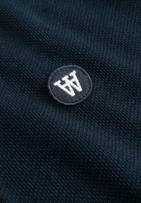 Tumši zila auduma virsma izceļas ar teksturētu austu struktūru, kurai centrā ir aplī izšūts logo jūras zilā un baltā krāsā ar burtiem "WW".
