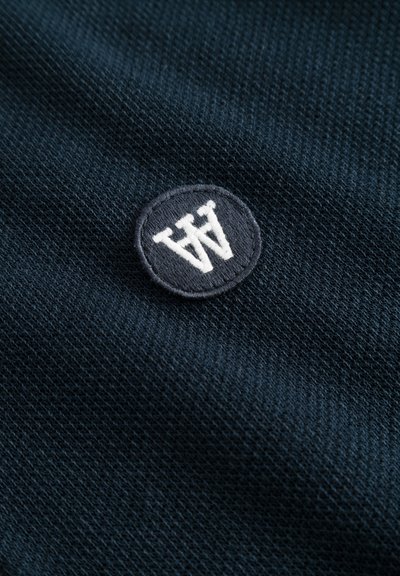 Un tessuto blu scuro presenta una trama testurizzata con un logo ricamato circolare in blu navy e bianco che mostra le lettere "WW."