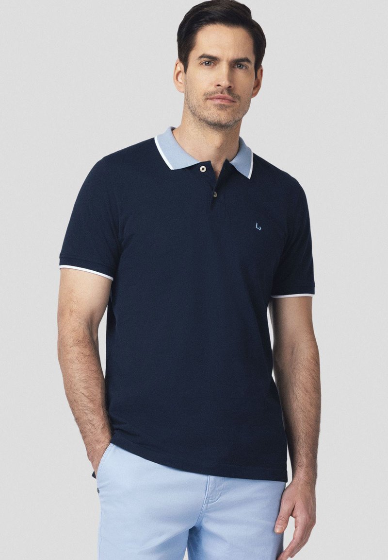 Granatowa koszulka polo z jasnoniebieskim kołnierzykiem, krótkimi rękawami i białymi akcentami. Posiada małe logo na lewej piersi.