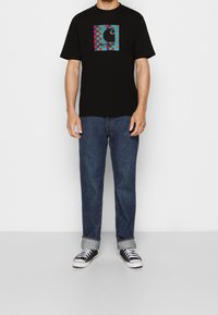 Camiseta negra con un gráfico de tablero de ajedrez colorido que presenta un logo. Combinada con jeans azules y zapatillas negras con suelas blancas.