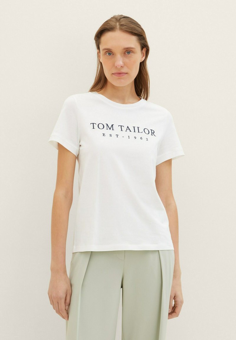 Fehér pamut póló rövid ujjakkal, középen sötétkék "TOM TAILOR EST. 1962" felirattal. Világoszöld nadrággal párosítva.