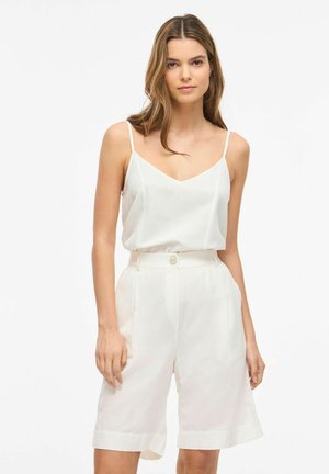 Mlada žena nosi bijelu majicu bez rukava s V-izrezom i bijele visokog struka kratke hlače do koljena, stoji ispred jednobojne pozadine.