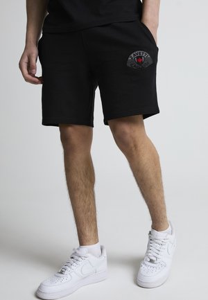 Mann iført svarte shorts med Zavetti-logo på lommen og hvite joggesko, stående mot en lys bakgrunn.