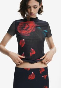 Schwarzes Blusenshirt mit einem großen roten Rosenmuster, aus locker sitzendem, transparentem Material und gerüschten Kanten. Wird mit einem passenden schwarzen Rock kombiniert.