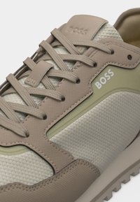 Gros plan d'une basket BOSS beige et crème avec un tissu texturé, des surpiqûres synthétiques, des lacets et le logo de la marque sur le côté et la languette.