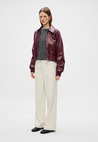 Κροπ δερμάτινο μπουφάν σε μπορντό απόχρωση με γυαλιστερή φινίρισμα, γκρίζα ζακέτα, κρεμ παντελόνι σε φάρδυ μέγεθος και μαύρα παπούτσια. Το μοντέλο στέκεται μπροστά σε έναν απλό φόντο.