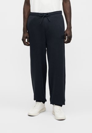 Άνδρας φοράει φαρδιά μαύρη φόρμα με κορδόνι στη μέση και λευκά αθλητικά, στέκεται μπροστά σε απλό φόντο.