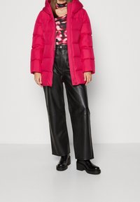 Chaqueta acolchada de color rosa brillante con capucha, diseño acolchado y cremallera. Combinada con pantalones anchos de cuero sintético negro y botas negras gruesas.