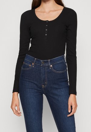 Frau trägt ein eng anliegendes schwarzes, geripptes Langarmshirt mit Knöpfen und hoch taillierte blaune Jeans vor neutralem Hintergrund.