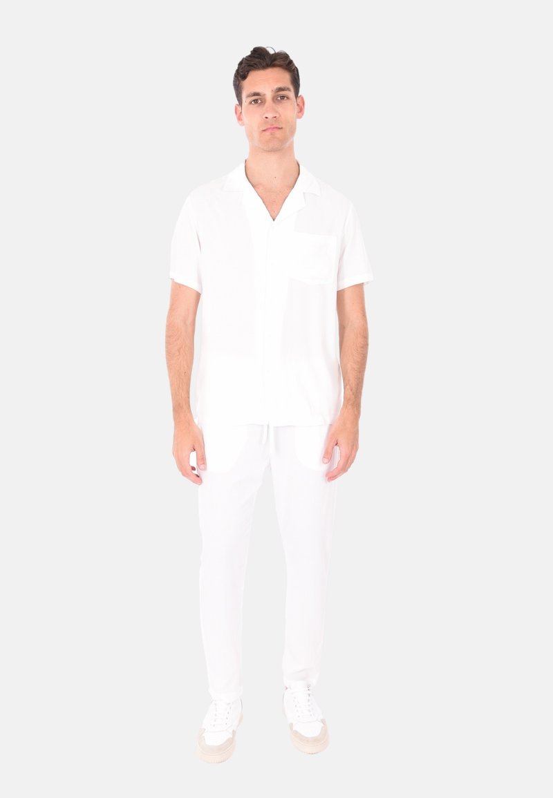 Camicia bianca a maniche corte con taschino, abbinata a pantaloni bianchi coordinati e sneakers bianche, il tutto in tessuto leggero.