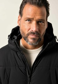 Schwarze Daunenjacke mit Reißverschluss, Kapuze und dezent gesteppten Einsätzen. Getragen über einem hellen, strukturierten Pullover.