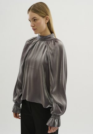 Blusa grigia chiaro in seta, con collo alto, dettagli plissettati e polsini arricciati e volant. Vestibilità morbida, texture liscia.