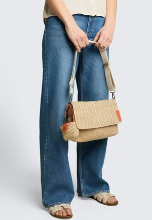 Osoba nosząca niebieskie dżinsy i beżowe sandały, trzymająca plecioną beżową torebkę na ramię z pomarańczowymi akcentami za pasek.