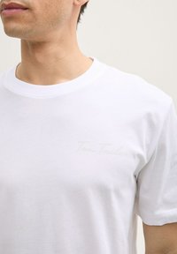 Hvid bomulds T-shirt med rund halsudskæring, korte ærmer og et diskret præget logo på brystet. Glat tekstur.