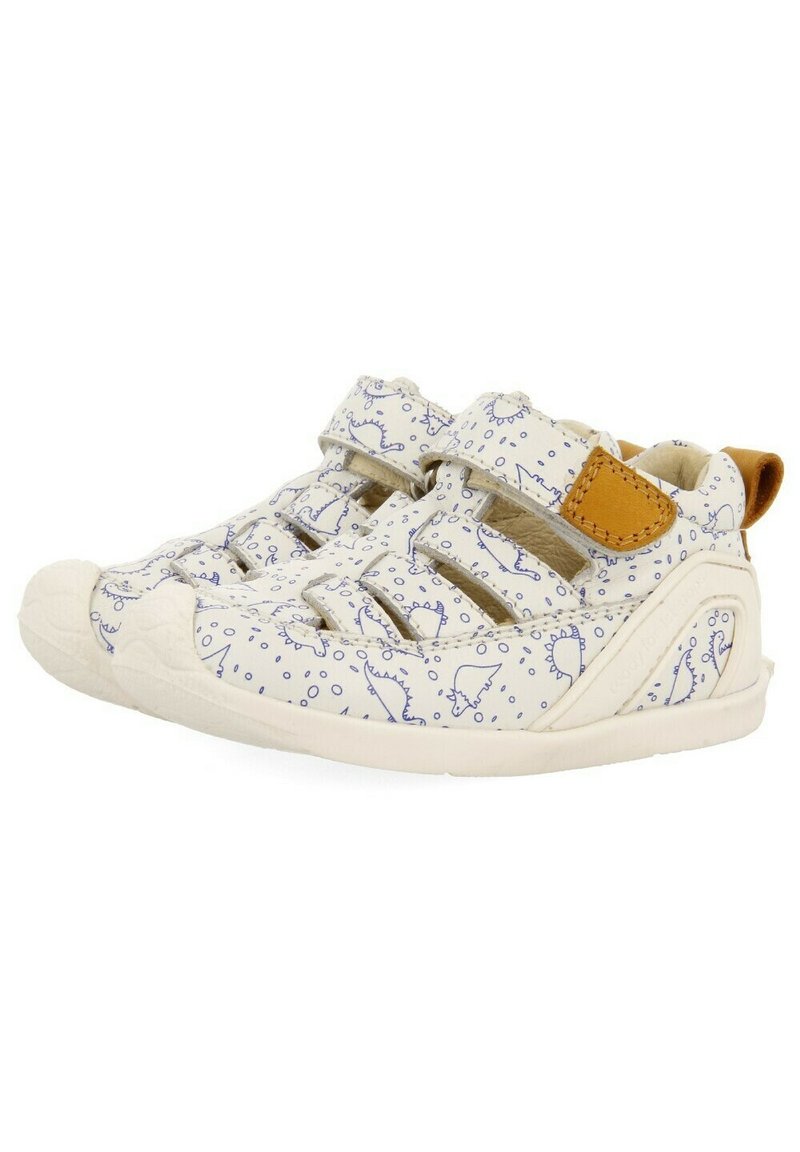 Zapatos blancos para niños con patrones de dinosaurios azules, asegurados con correas de Velcro, puntera redondeada y un acento de cuero marrón en el talón.