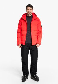 Rote Daunenjacke mit Kapuze, ausgestattet mit gesteppten Einsätzen. Tragen über einem schwarzen Langarmshirt und kombiniert mit schwarzen Hosen und Stiefeln.