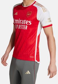 Red football jersey with a zigzag texture, white sleeves, and gold accents. Logos for Emirates and Arsenal displayed prominently.