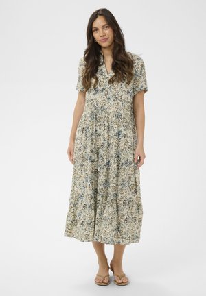 Woman with long brown hair wearing a short-sleeve, floral-patterned midi dress and beige sandals, standing against a plain white background.