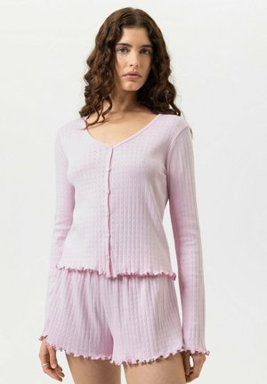 Vrouw met krullend haar, gekleed in een lichtroze geribbelde lange mouwen top met knopen en bijpassende korte broek met gerimpelde randen, staand tegen een witte achtergrond.