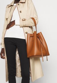 Brun läderhinkväska med snörstängning, slät textur och guldfärgade detaljer. Bärs med en beige trenchcoat och svarta byxor.