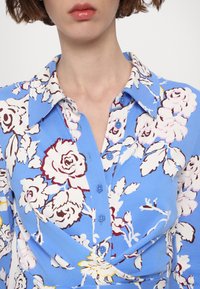 Kék virágmintás blúz gombos elülső résszel, nagy gallérral, valamint kontrasztos fehér és rózsaszín rózsákkal, zöld levelekkel.