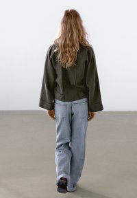 Giacca verde scuro con maniche larghe, abbinata a jeans in denim azzurro. Modello visto da dietro su uno sfondo neutro.