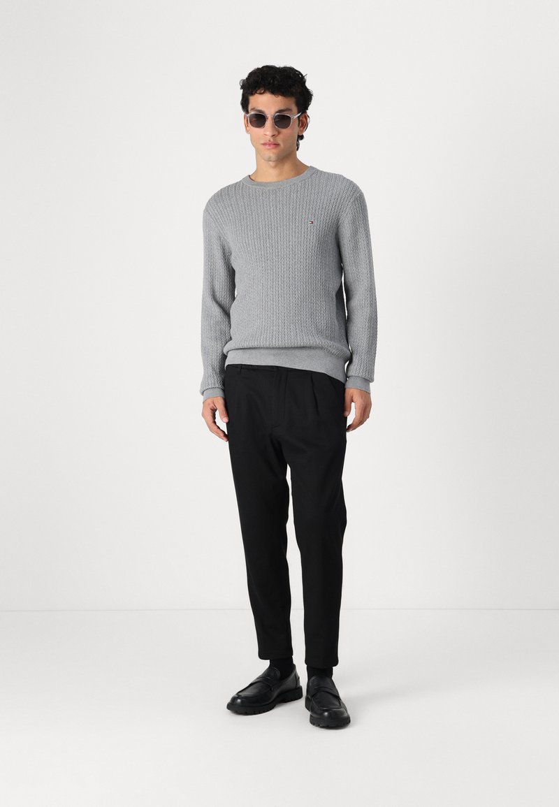 Pull en tricot gris avec un motif texturé, associé à un pantalon noir et des chaussures noires. Les accessoires incluent des lunettes de soleil rondes.