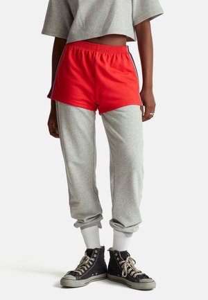 Personne portant un short de sport rouge par-dessus un pantalon de survêtement gris, des chaussettes blanches, des baskets montantes noires et un T-shirt gris court.
