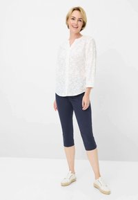 Blusa bianca ricamata con bottoni frontali e maniche a tre quarti, abbinata a pantaloni capri blu navy e sneakers bianche con dettagli in juta.