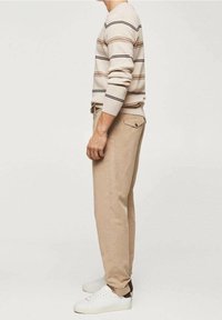 Maglione collaborativo beige con righe orizzontali marroni, nere e rosa, abbinato a pantaloni beige chiari e sneakers bianche.