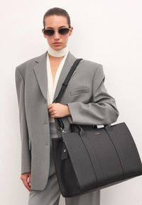 Fekete textúrált bőr tote táska dupla felső fogantyúval, vállpánttal és egy kis címkével. Szürke, túlságosan nagy zakóval viselve.