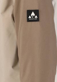 Beige stofjakke ærme med en sort firkantet lap med et hvidt bjerglogo og teksten "WHISTLER". Glat tekstur, skræddersyet pasform.