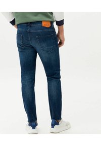 Blue denim jeans with a slim fit, featuring a brown leather brand patch at the back, and two rear pockets. Worn with white sneakers.