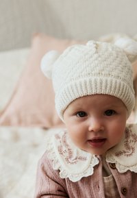 Cappellino bianco di lana per neonati con pon-pon, abbinato a un cardigan rosa con trama e un colletto color crema con ricami floreali.