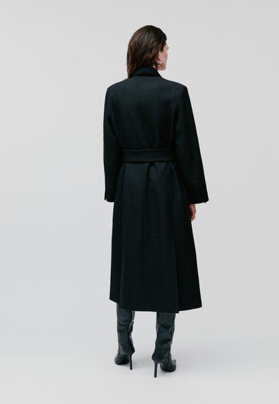 Cappotto lungo nero con vita a cintura, ampio colletto e maniche lunghe. Tessuto liscio con silhouette sartoriale, abbinato a stivali neri sopra il ginocchio.