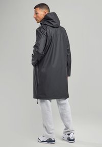 Black waterproof coat with a hood, knee-length design, and drawstring adjustment. Worn over gray sweatpants and white sneakers.