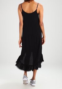 Musta ilman hihoja mekko, jossa on röyhelöhelma, takanäkymä. Valmistettu kevyestä kankaasta, ohuiden olkaimien ja hulmuavan siluetin kanssa.