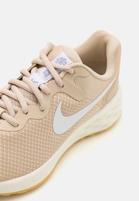 Zapatilla de running Nike beige con swoosh blanco, parte superior de malla, suela acolchada y lazada beige sobre un fondo blanco.